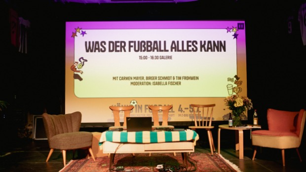 Foto von der Bühne im Z-Bau in Nürnberg bei der Veranstaltung "König*in Fußball". Zu sehen ist die noch leere Bühne mit Aufbau für ein Podiumsgespräch. Auf der Leinwand ist zu lesen "Was der Fußball alles kann"