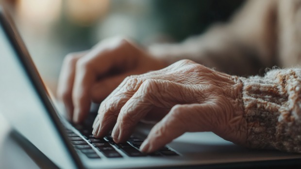 Nahaufnahme der faltigen Hände einer älteren Person beim Tippen auf einer Laptop-Tastatur